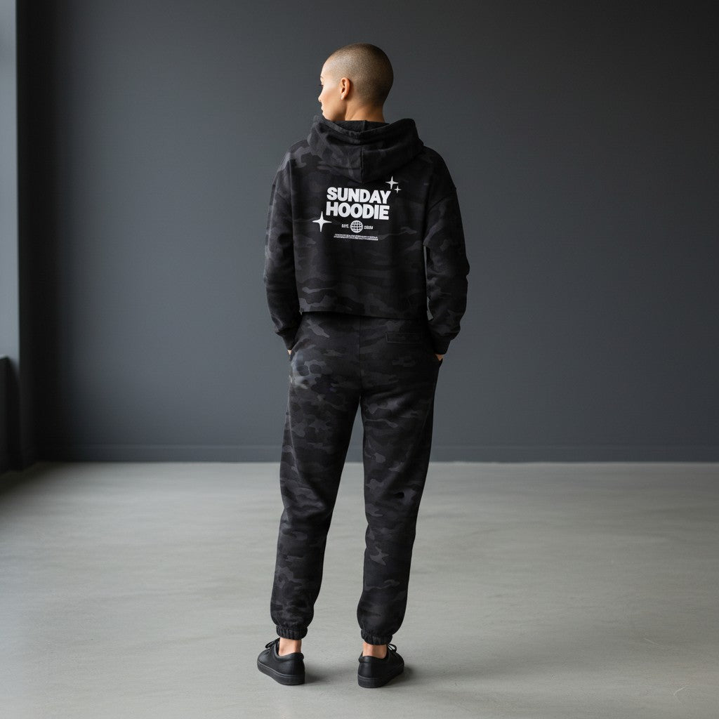 Model wearing Omina black camo cropped Sunday hoodie and matching joggers from behind in a minimalist studio.