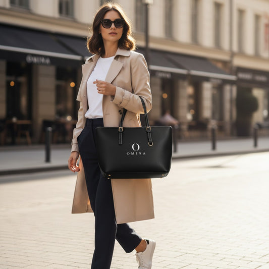 Woman in trench coat carrying a black Omina PU leather shoulder tote bag with gold hardware on a city street.