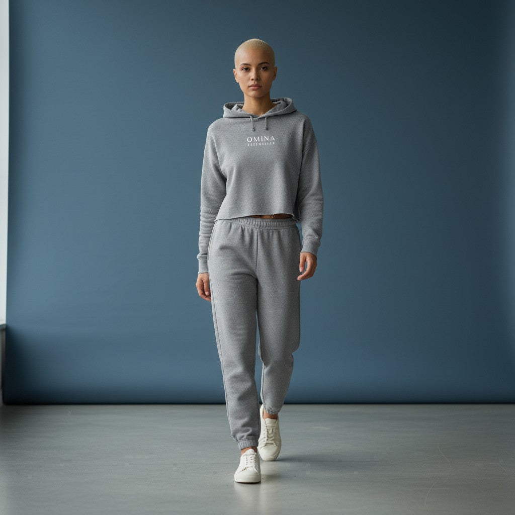 Model wearing Omina Essentials grey cropped hoodie and matching fleece joggers against a blue studio backdrop.
