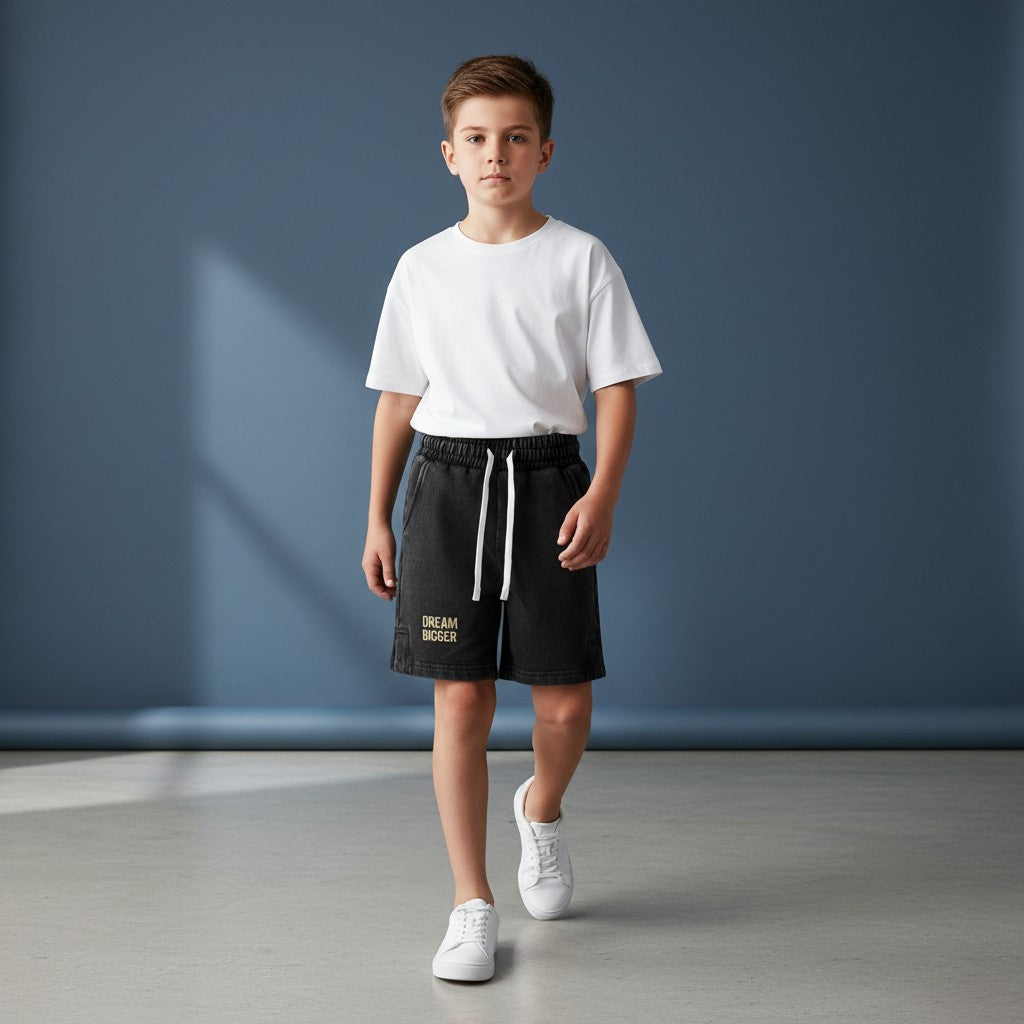 Omina Kids snow-washed black shorts with elastic waistband, white drawstring, and "DREAM BIGGER" graphic.