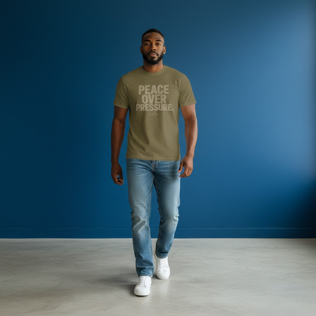 Model wearing an Omina olive green heavyweight cotton t-shirt with PEACE OVER PRESSURE text against a blue wall.