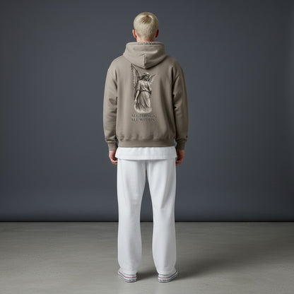 Omina Protected By Grace oversized taupe hoodie with angel graphic on back worn by a model in white pants.