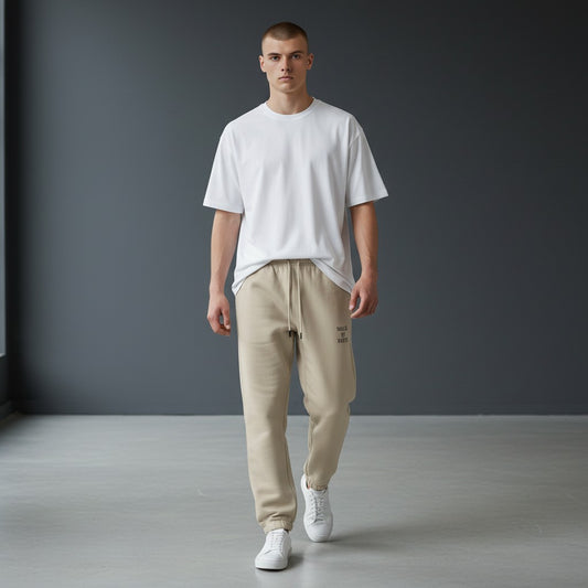 Omina Studio beige cotton sweatpants with "WALK BY FAITH" embroidered on the left thigh. Features elastic waistband, drawstring, and tapered legs.