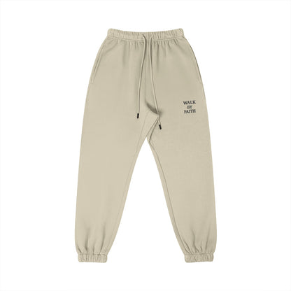 Omina Studio beige cotton sweatpants with "WALK BY FAITH" embroidered on the left thigh. Features an elastic waistband, drawstring, and tapered leg.
