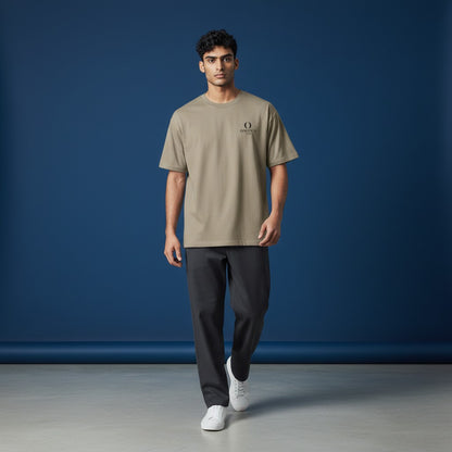 Model wearing an Omina heavyweight taupe cotton t-shirt with a small chest logo, paired with dark trousers.